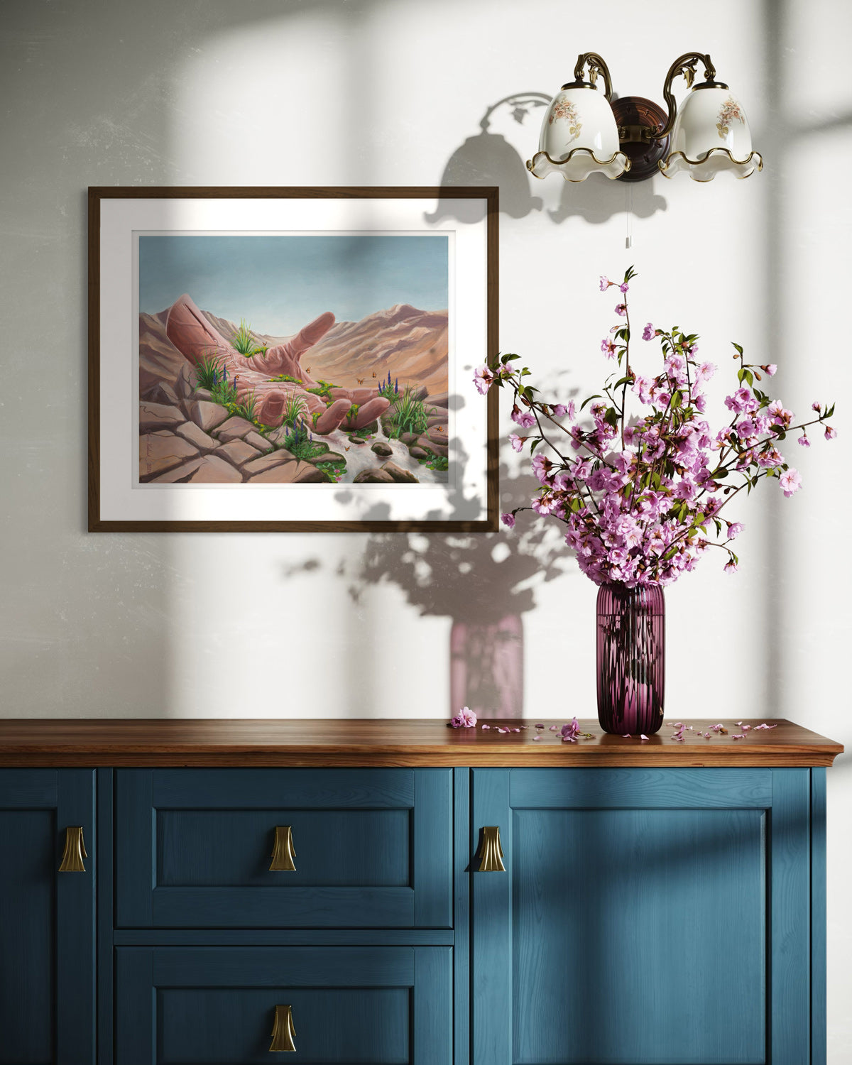 A framed print of the artwork titled 'Calm Surrender' by Glenn Verkerk, created with acrylics on linen canvas. The artwork depicts an open hand resting in a dry desert, with a small stream of water flowing from the wrist, creating a lush, green landscape with small waterfalls, streams, butterflies, and flowers. The artwork is hanging on the wall above a country side cabinet with pink flowers and a lamp.