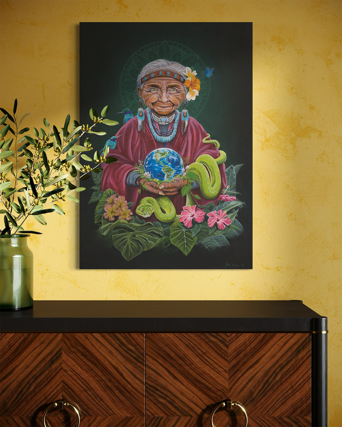 Designer cabinet, olive tree branches, and a yellow wall featuring the original canvas titled 'Pachamama' by Glenn Verkerk. The artwork depicts an imaginative version of Mother Earth holding planet Earth, inspired by Inca tribe traditions and symbolism. The scene is surrounded by a mandala, animals, and lush green elements.