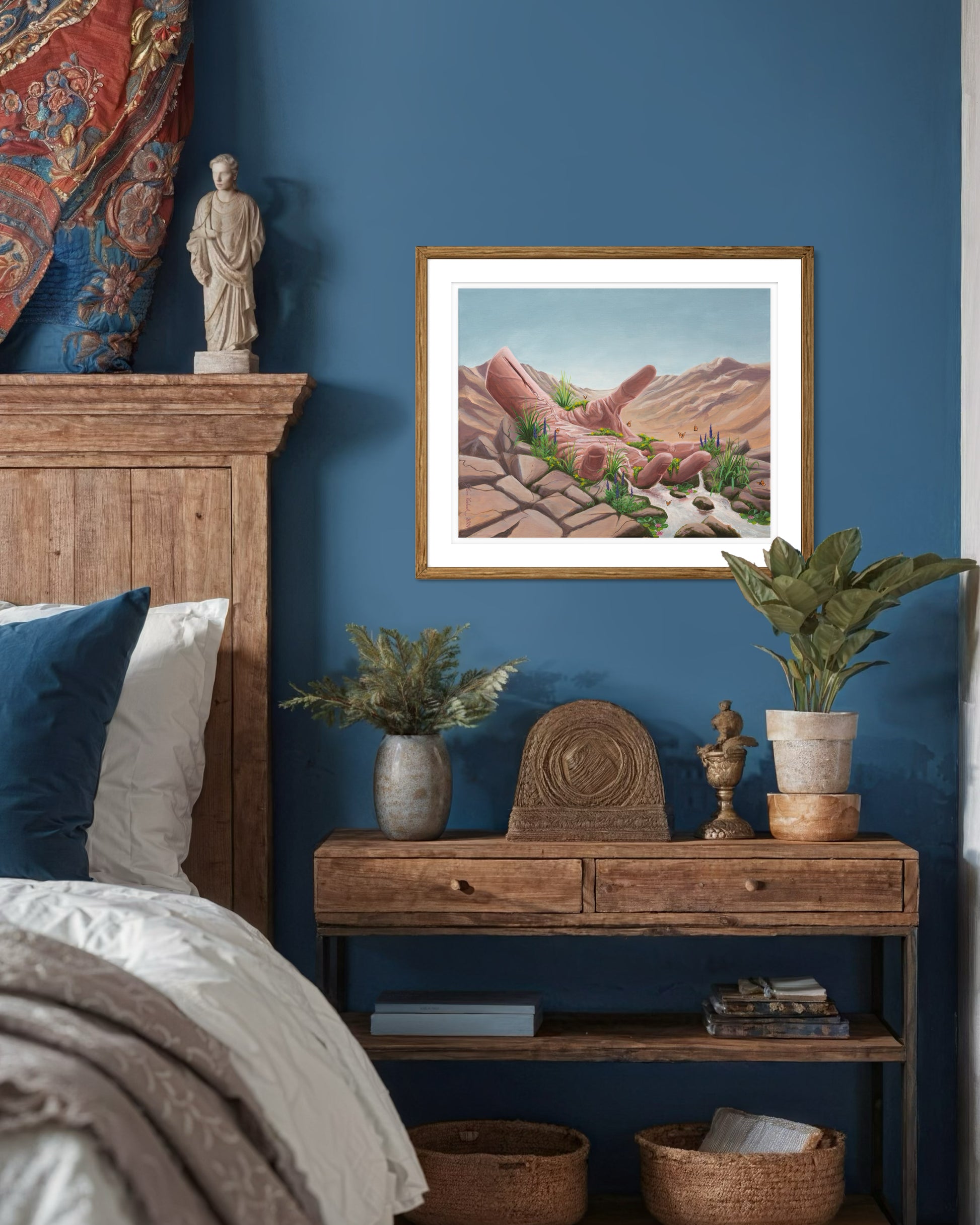 A framed print of the artwork titled 'Calm Surrender' by Glenn Verkerk, created with acrylics on linen canvas. The artwork depicts an open hand resting in a dry desert, with a small stream of water flowing from the wrist, creating a lush, green landscape with small waterfalls, streams, butterflies, and flowers. The artwork is hanging on a blue wall above a nightstand in a stylish bedroom. 