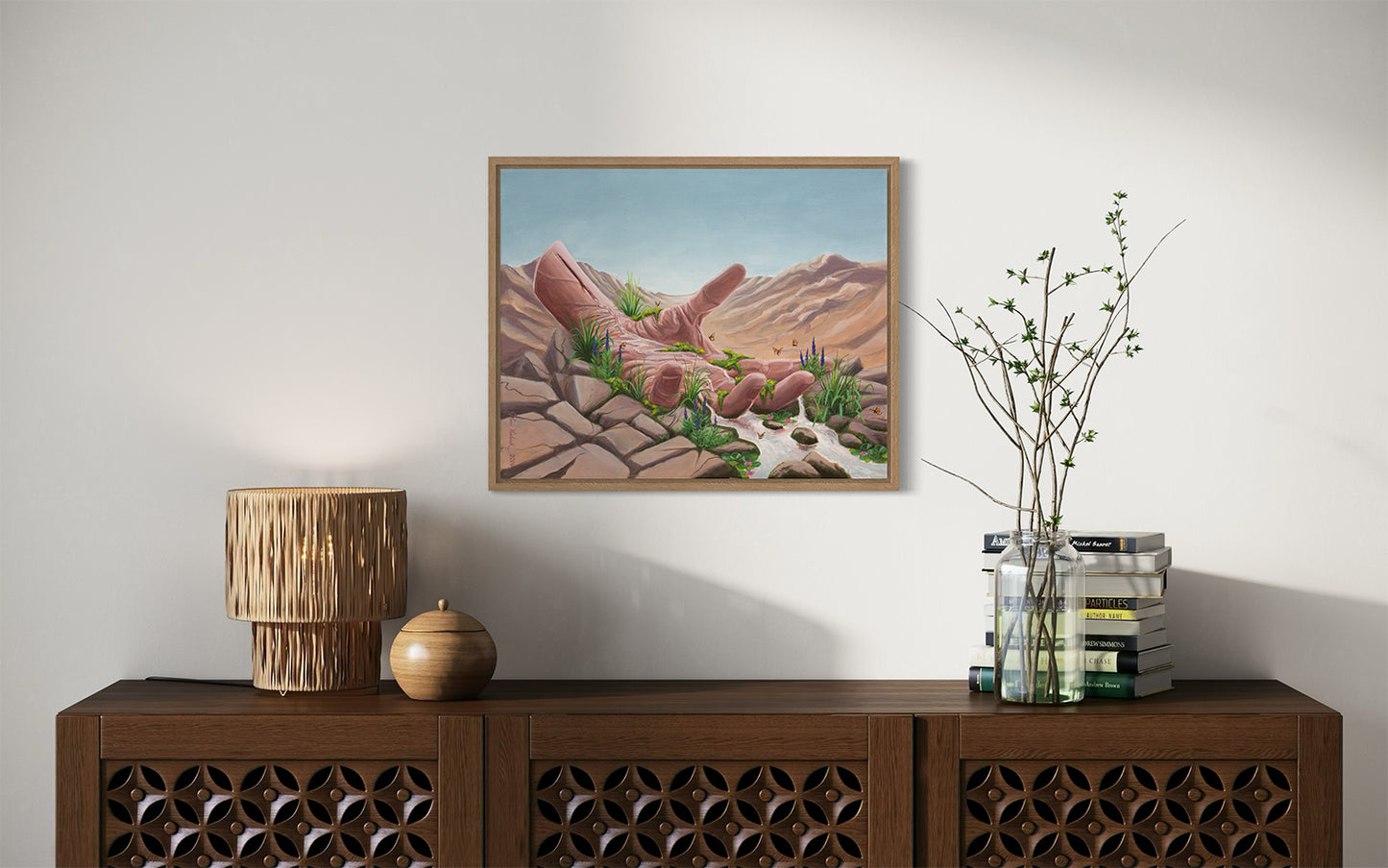 The original artwork titled 'Calm Surrender' by Glenn Verkerk, created with acrylics on linen canvas. The artwork depicts an open hand resting in a dry desert, with a small stream of water flowing from the wrist, creating a lush, green landscape with small waterfalls, streams, butterflies, and flowers. The artwork is hanging on the wall above a stylish cabinet with some books and a lamp.